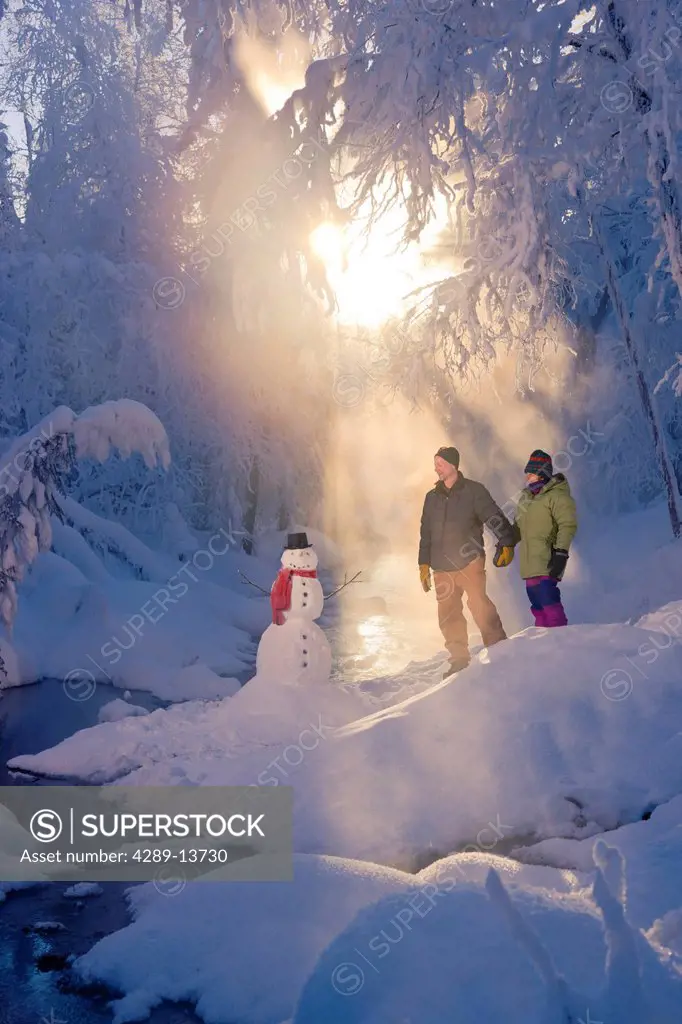 Husband and wife standing in a frosty forest next to a snowman and backlit by sunrays, Russian Jack Springs Park, Southcentral Alaska, Winter