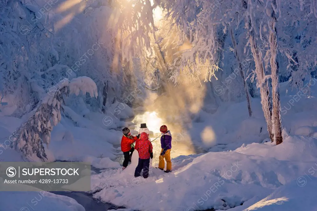 Three children building a snowman next to a stream in a frost covered forest, Russian Jack Springs Park, Anchorage, Southcentral Alaska, Winter