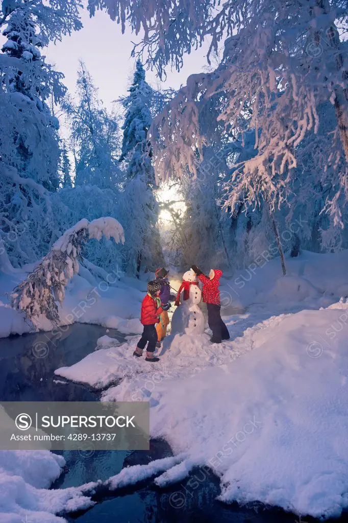 Three children building a snowman next to a stream in a frost covered forest, Russian Jack Springs Park, Anchorage, Southcentral Alaska, Winter