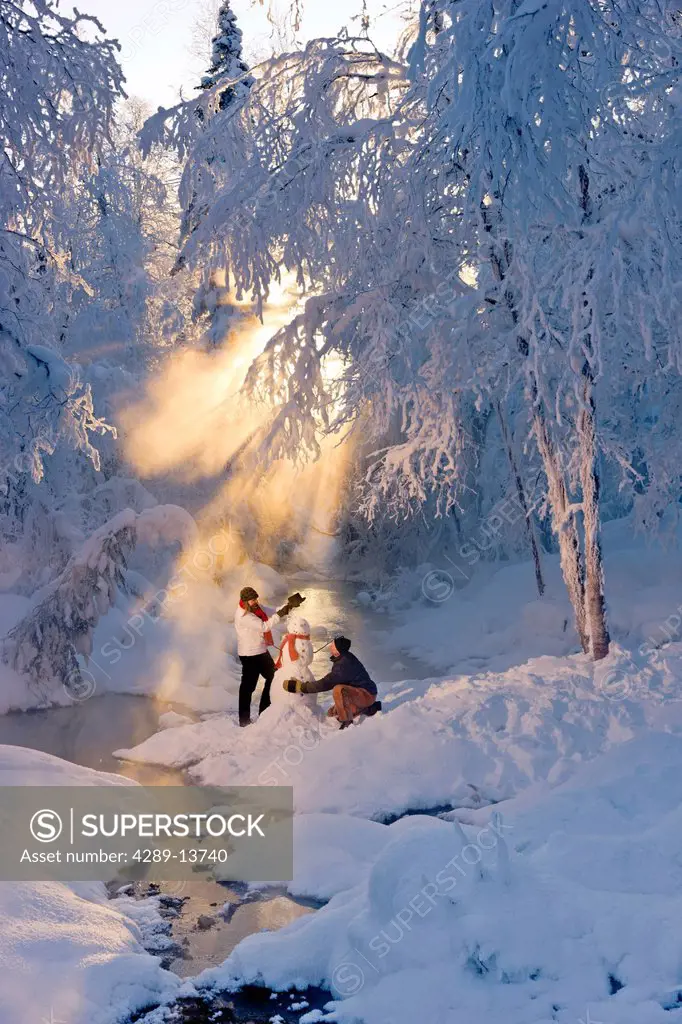 Husband and wife building a snowman in a frosty forest and backlit by sunrays, Russian Jack Springs Park, Southcentral Alaska, Winter