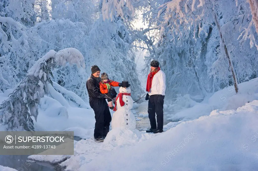 Man holding up his daughter to put a hat on a snowman while his wife watches with frost covered trees in the background, Russian Jack Springs Park, An...