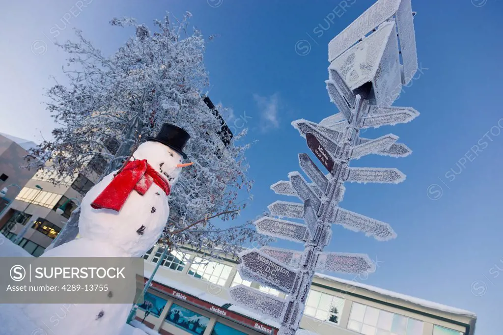 Snowman wearing a black top hat and red scarf standing next to the mileage signpost by the visitors log cabin in downtown Anchorage, Southcentral Alas...