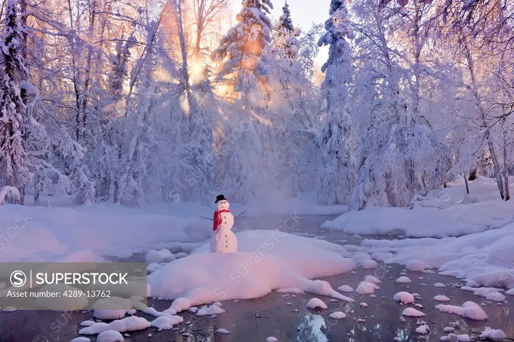 Snowman standing on a small island in the middle of a stream with sunrays shining through fog and hoar frosted trees in the background, Russian Jack S...