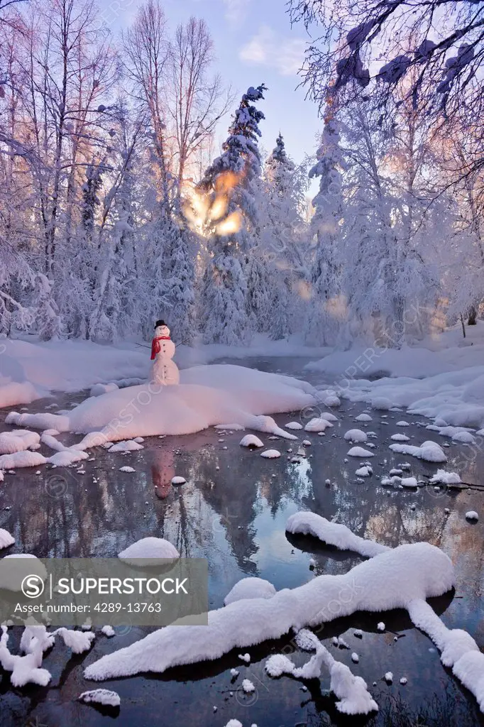 Snowman standing on a small island in the middle of a stream with sunrays shining through fog and hoar frosted trees in the background, Russian Jack S...