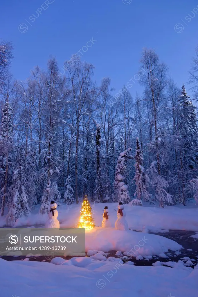 Snowman family standing next to a Christmas tree on a snow covered island in the middle of a small stream in a hoarfrost covered forest at twilight, R...