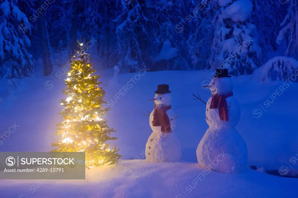 Two snowmen standing next to a Christmas tree on a snow covered island in the middle of a small stream in a hoarfrost covered forest at twilight, Russ...