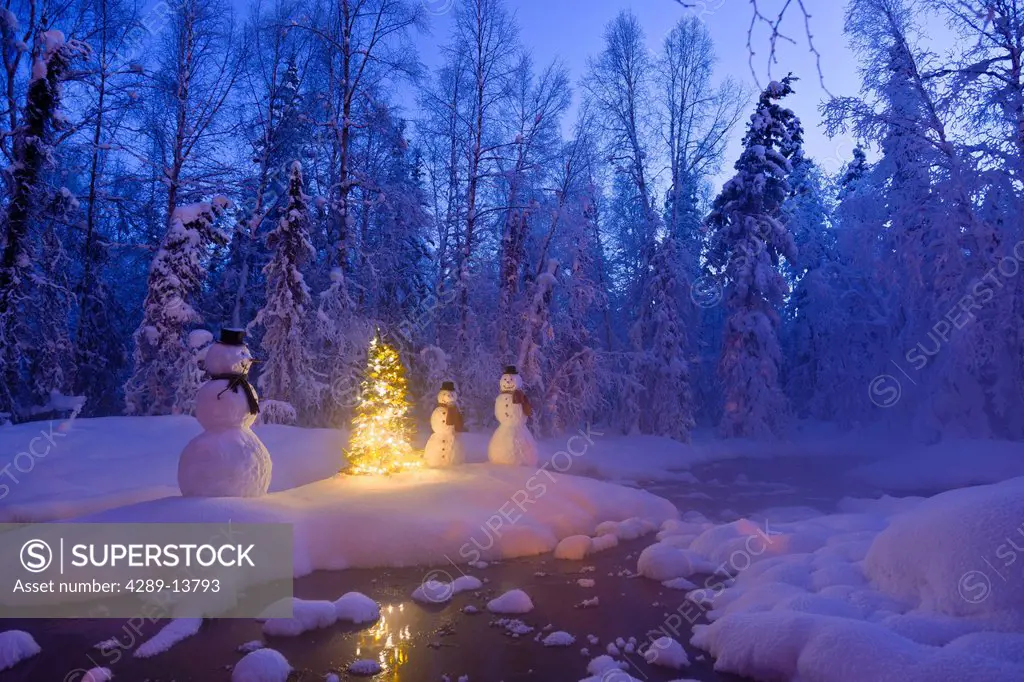 Snowman family standing next to a Christmas tree on a snow covered island in the middle of a small stream in a hoarfrost covered forest at twilight, R...