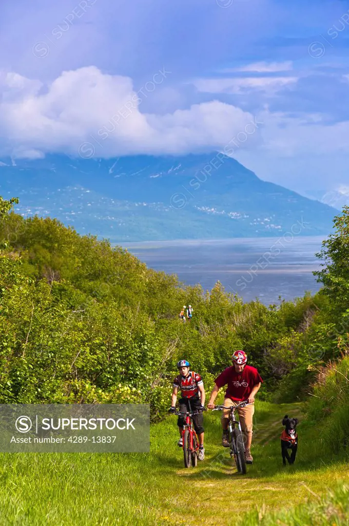 Two men mountain biking with their dog on a trail in Kincaid Park with a view of Cook Inlet in the background, Anchorage, Southcentral Alaska, Summer