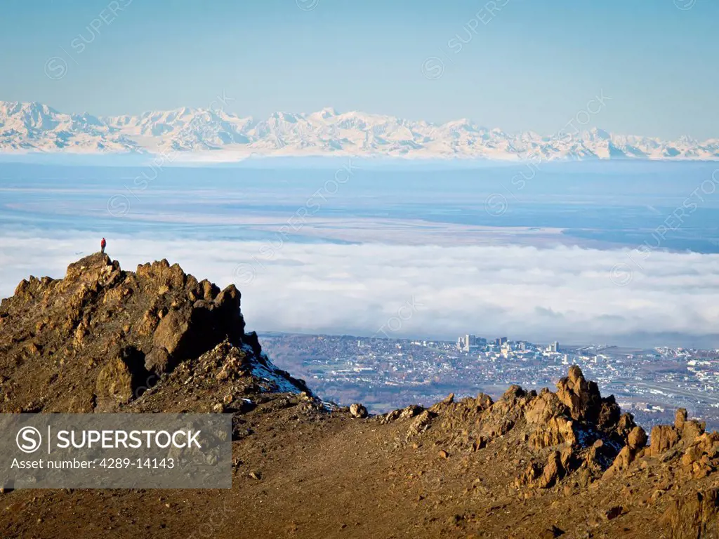 Woman trail runner stands on a rock ridge high above Anchorage with the Alaska Range beyond, Chugach Mountains, Southcentral Alaska, Autumn