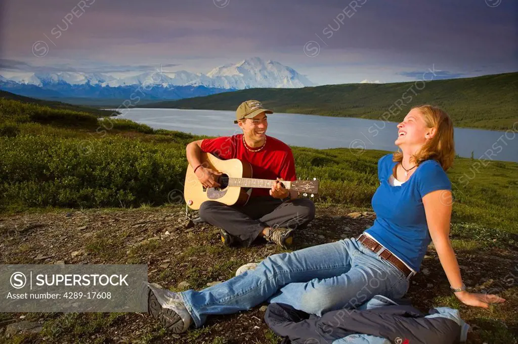 A couple sit on the tundra as the man plays guitar in Denali National Park with Wonder Lake in the background Alaska summer