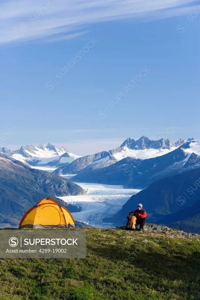 Hiker next to tent on ridge views Mendenhall Glacier & Coast Mountains near Juneau Alaska during Summer
