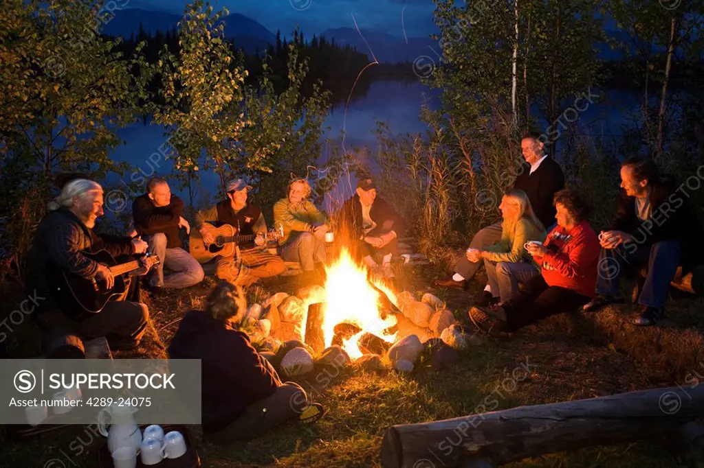 Group of adults sitting around a campfire sing and and talk as they enjoy the warmth of the fire and each others company at Winter Lake Lodge. Summer ...