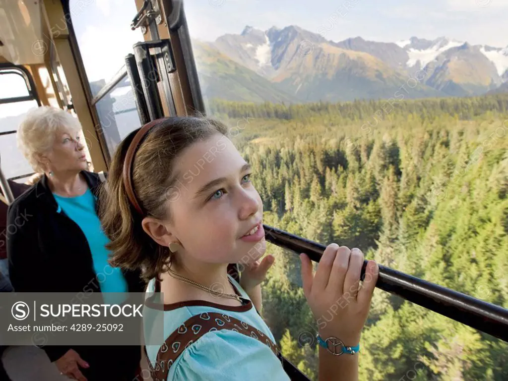 A girl rides with her grandmother up the Alyeska Resort tram with the scenic Girdwood bowl & Chugach Mountains in the background, Southcentral Alaska