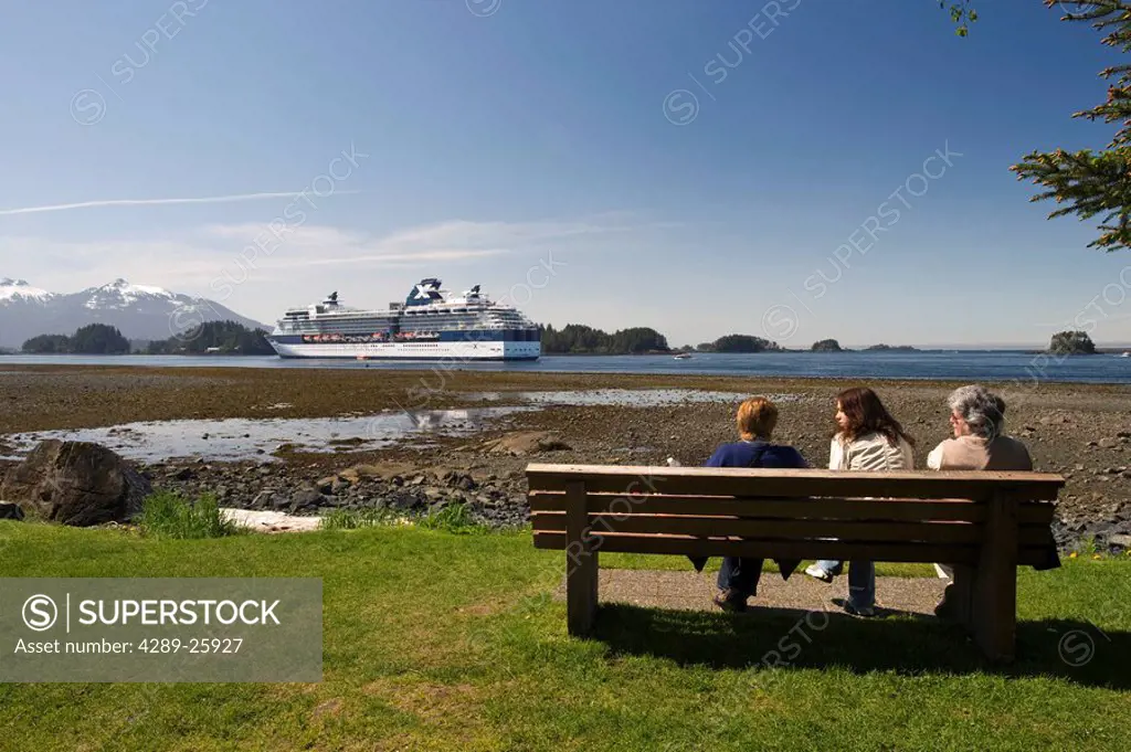 Tourists relax on a bench along the shore with a view of Celebrity cruise ship Infinity anchored at Sitka, Alaska