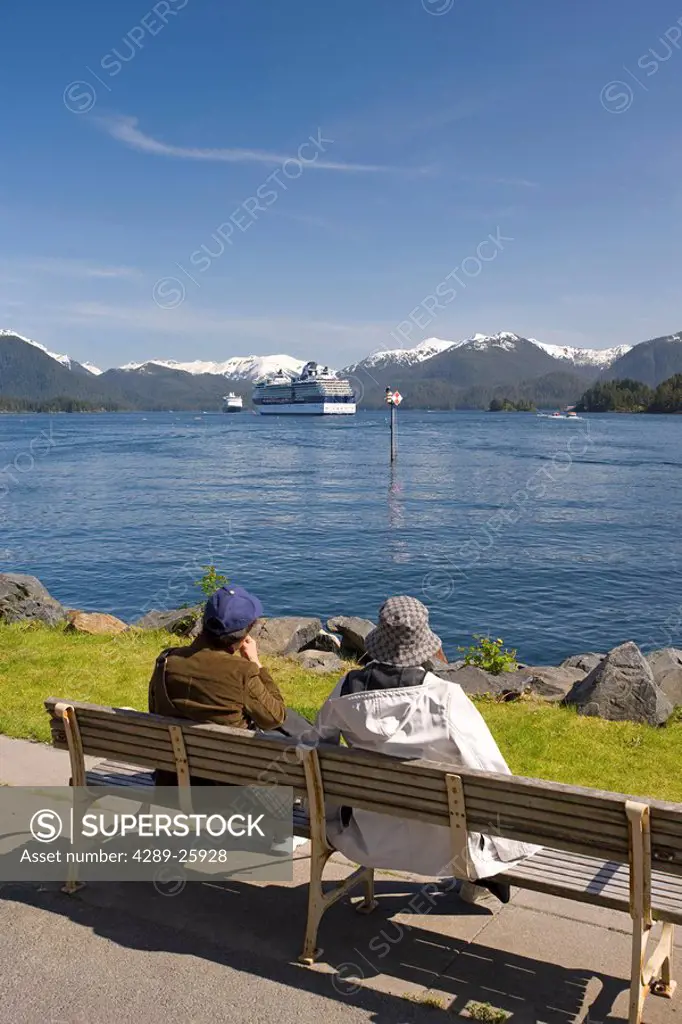 Tourists relax on a bench along the shore with a view of a Celebrity and a Holland America cruise ship anchored at Sitka, Alaska
