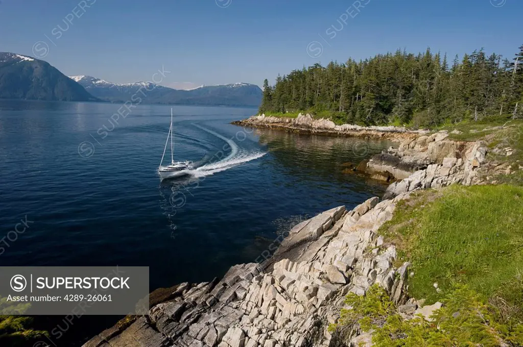 A motor powered sailboat runs along the shore of Perry Island in Prince William Sound on a sunny summer day in Southcentral Alaska