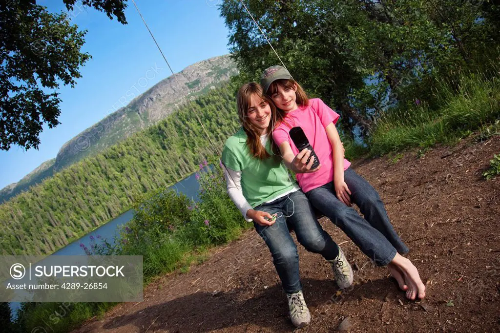 Two young girls sitting on a rope swing looking at photos on their cell phone, Byers Lake, Denali State Park, Southcentral Alaska