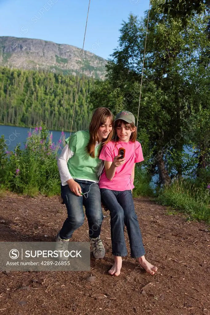 Two young girls sitting on a rope swing looking at photos on their cell phone, Byers Lake, Denali State Park, Southcentral Alaska