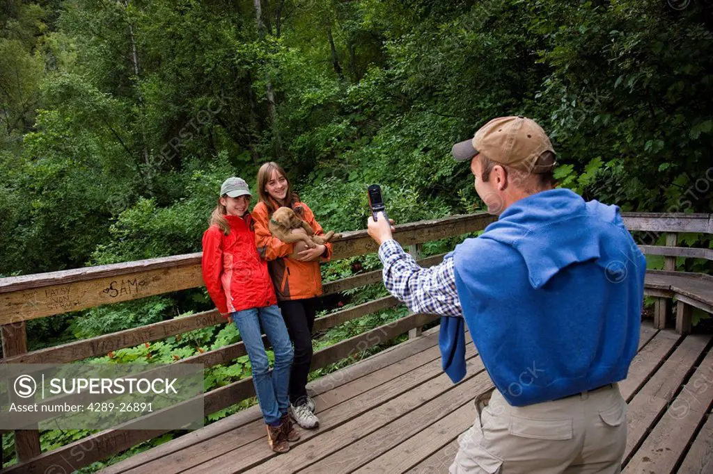 Father uses a cell phone to take a photo of his two teenaged daughters holding a puppy, Thunderbird Falls Overlook, Alaska