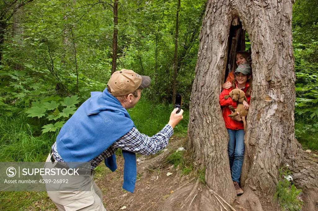 Father taking a picture of his two teenaged daughters standing inside a hollowed out Cotttenwood tree, holding a puppy, Thunderbird Falls, Eklutna, So...