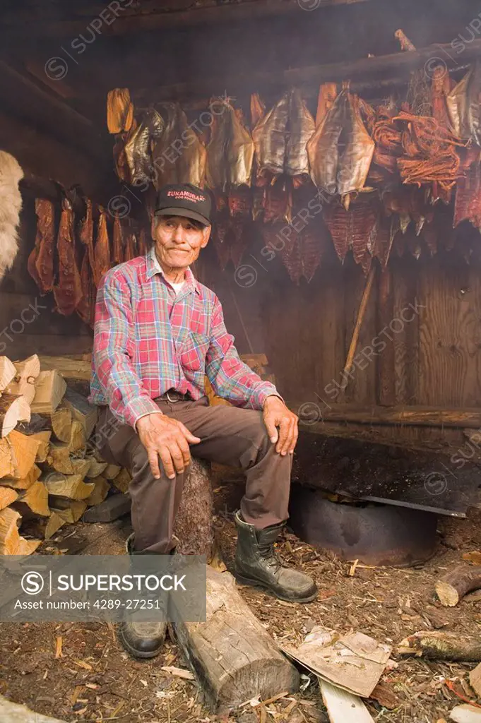Elder native Yupik man sitting in smokehouse w/hanging salmon tending fire Tuluksak WE Alaska