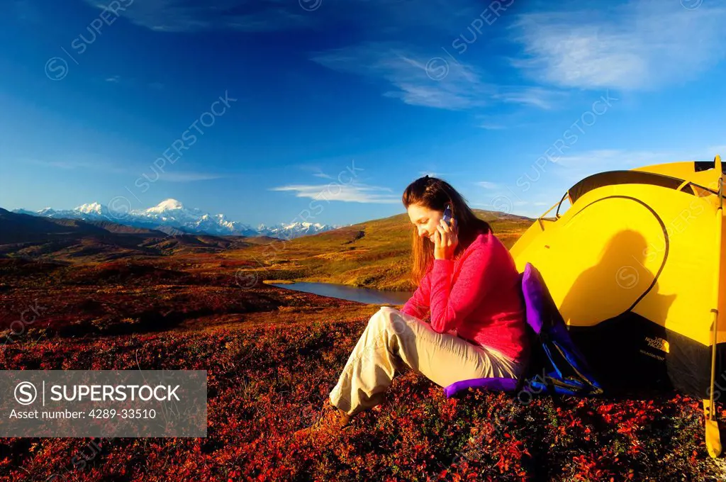 A woman talks on her cell phone and takes a picture of Mt. McKinley while relaxing by her tent in Peters Hills, Denali State Park, Southcentral Alaska...