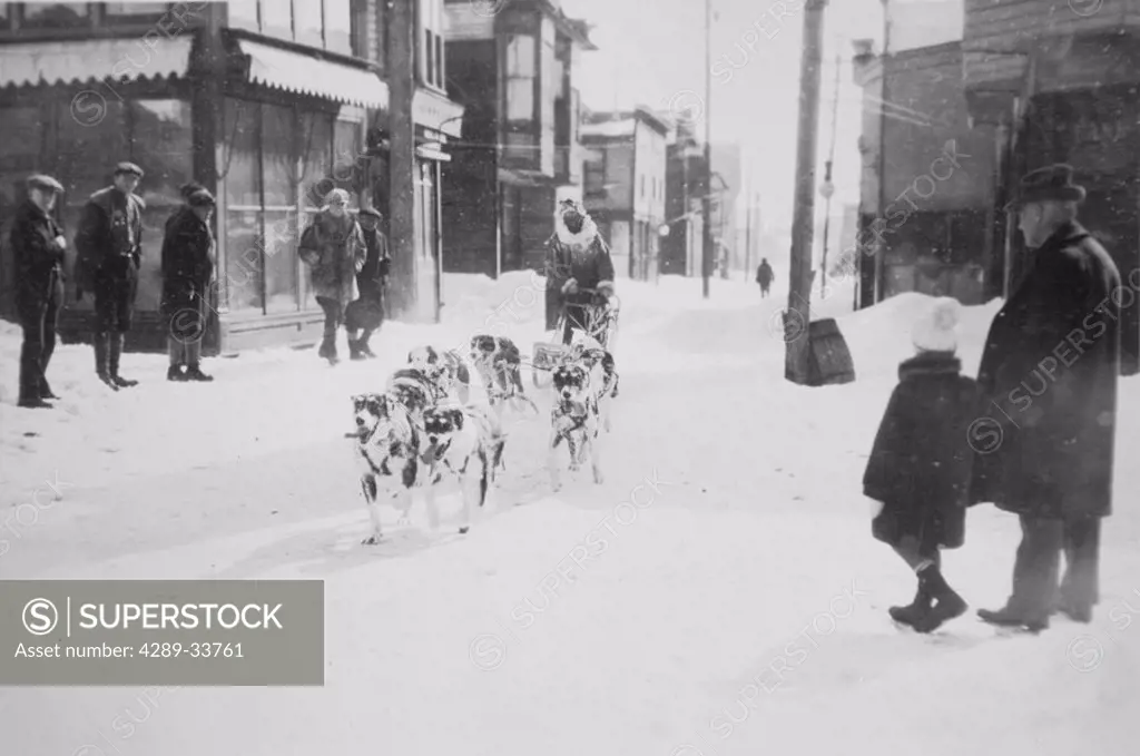 Historical image of sled dog team running down street in Nome and a few observers winter Nome WE Alaska