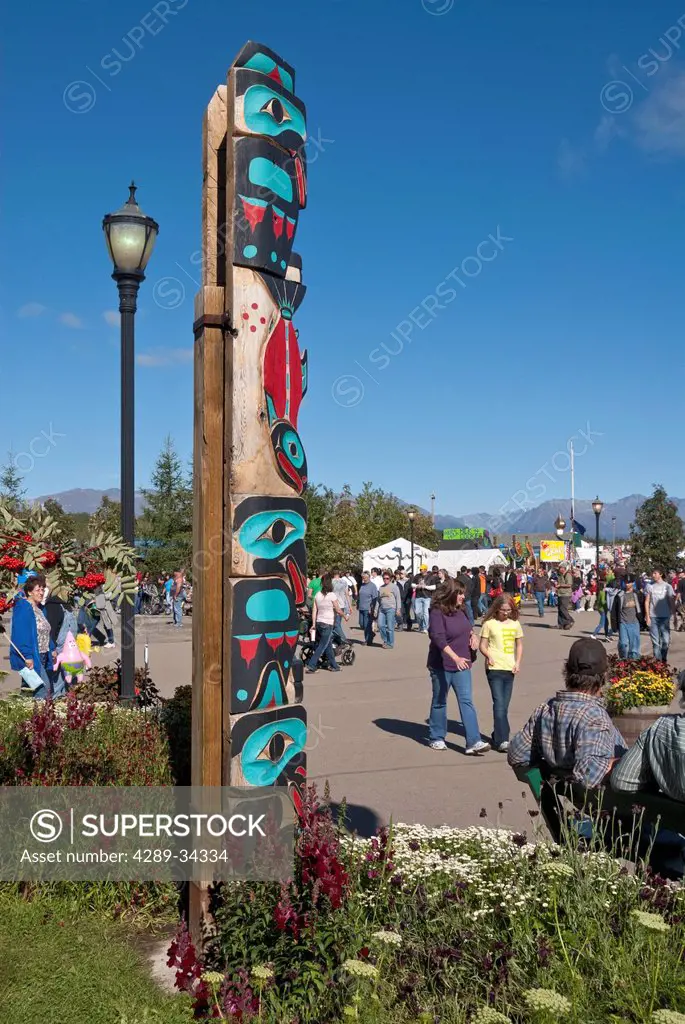 People walk by a totem pole near Raven Hall at Pioneer Plaza at the Alaska State Fair, Palmer, Southcentral Alaska, Autumn