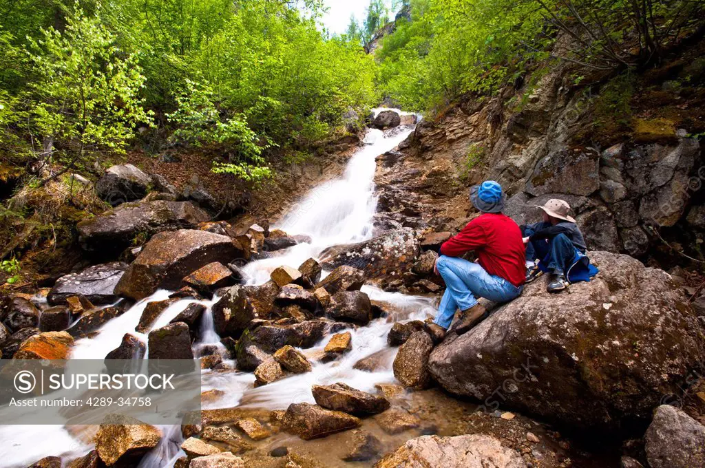 A father and son on a hike rest on a rock while looking at Cascade Falls near Byers Lake in Denali State Park, Southcentral Alaska, Summer