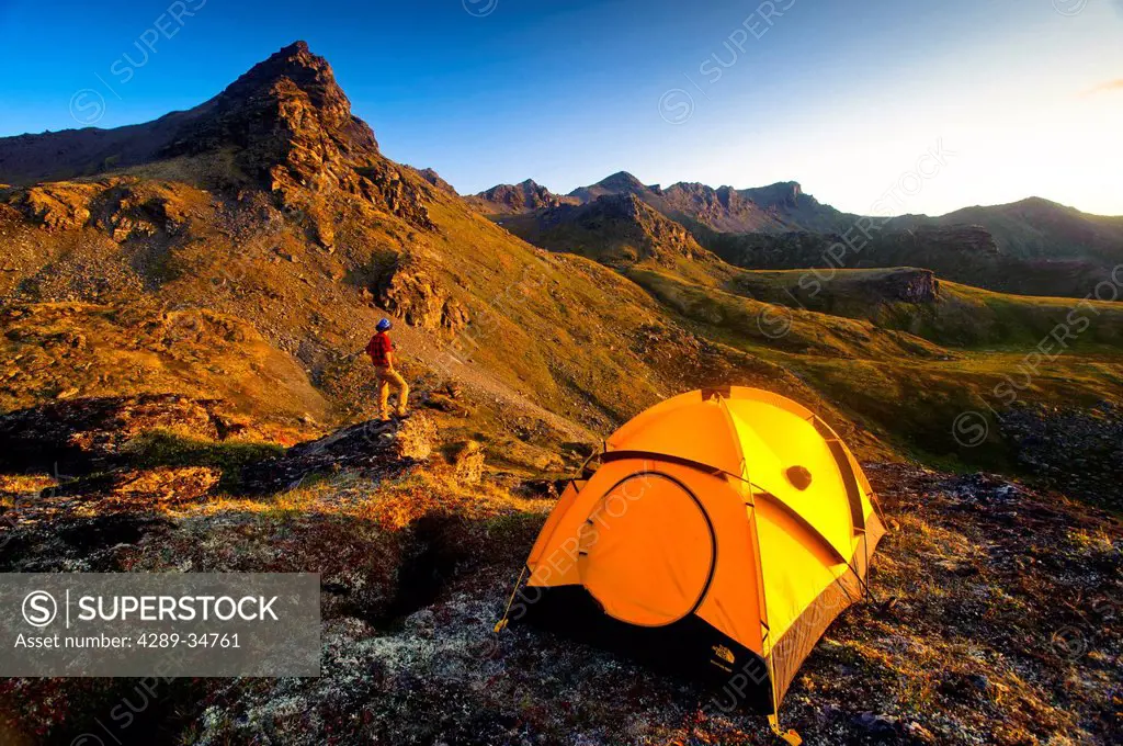 A man standing on a rock next to his tent while camping near Hatcher Pass in the Talkeetna Mountains with Bald Ridge in the background, Southcentral A...