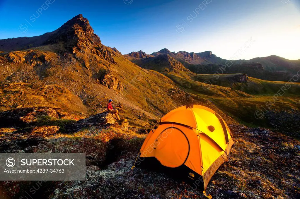 A man standing on a rock next to his tent while camping near Hatcher Pass in the Talkeetna Mountains with Bald Ridge in the background, Southcentral A...