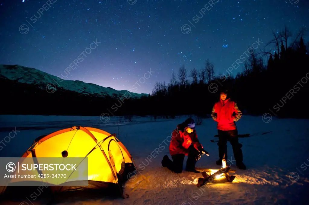 Two men warm themselves at a campfire while standing next to their lighted tent at their campsite on Eklutna Lake in the Chugach State Park, Southcent...