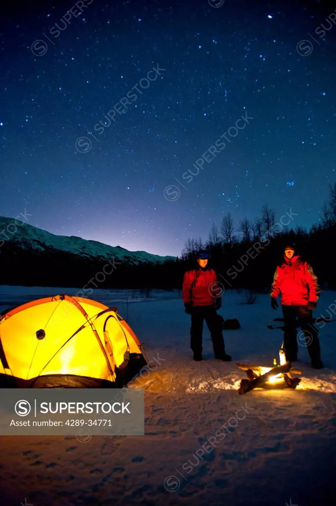 Two men warm themselves at a campfire while standing next to their lighted tent at their campsite on Eklutna Lake in the Chugach State Park, Southcent...
