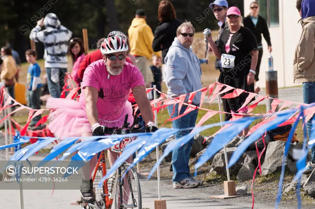 Senior man dressed in a pink shirt and tutu costume bikes across the finish line during the Pink Cheeks Triathlon, Seward, Southcentral Alaska, Summer