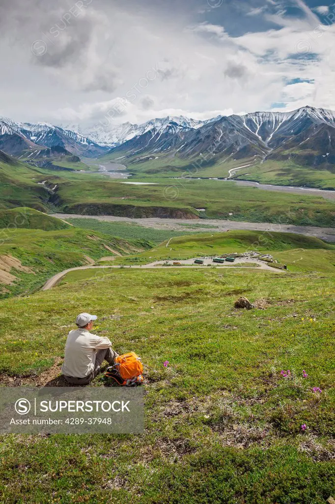 A Senior Male Hiker Sitting On The Tundra On The Mountain Side Looking Down At The Eielson Visitor's Center In Denali National Park; Alaska United Sta...