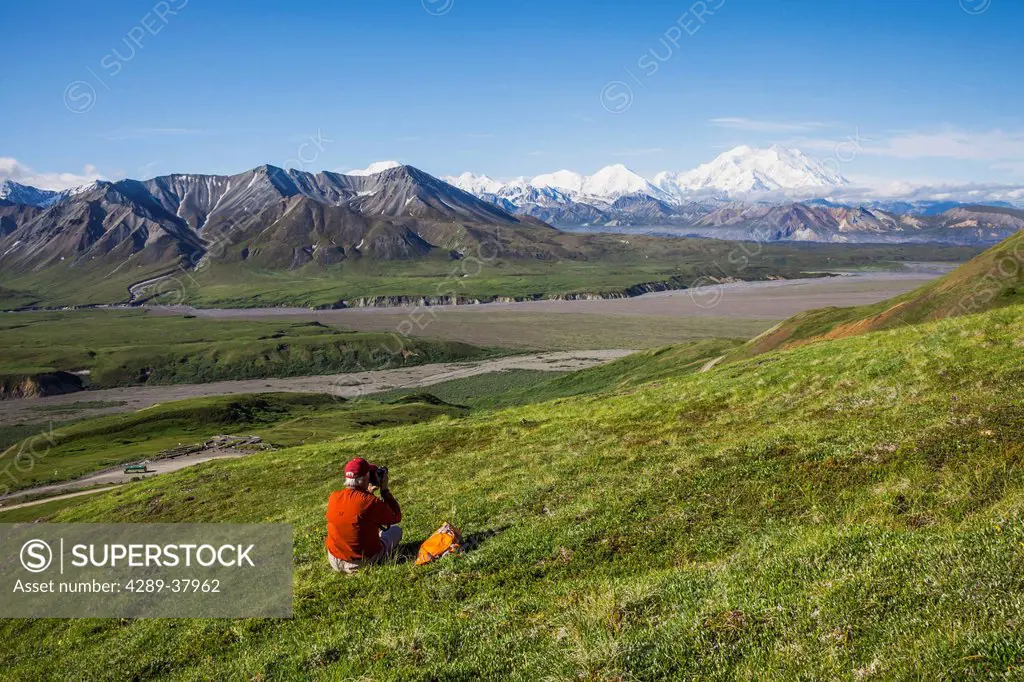 A Senior Male Hiker Sits On The Tundra On A Mountain Slope With Mt. Mckinley And The Thorofare River In The Background In Denali National Park; Alaska...