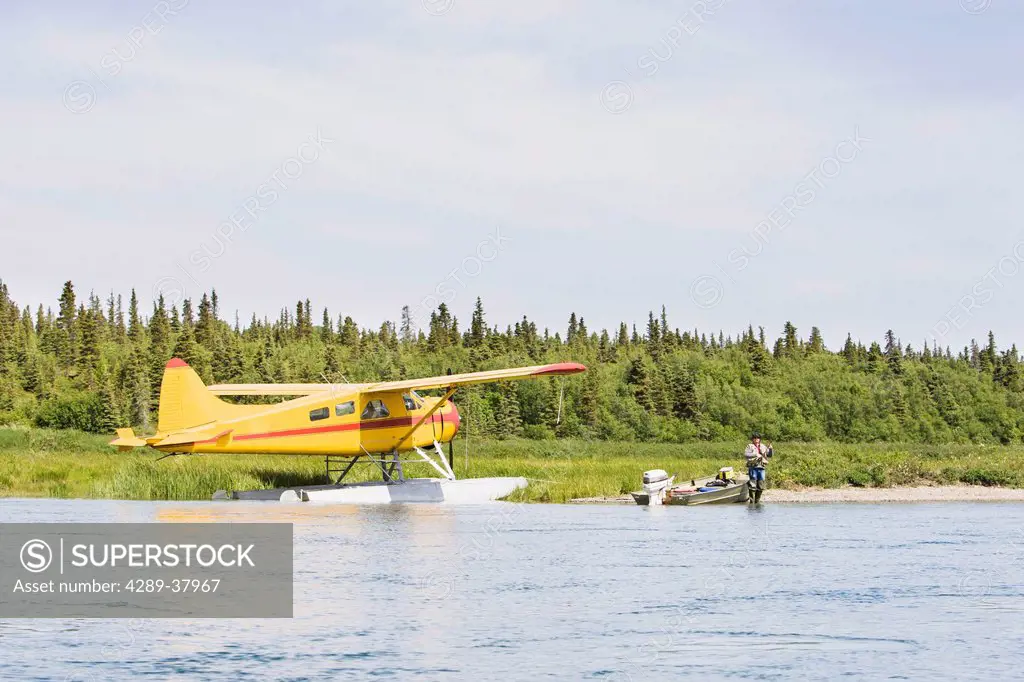 A Sport Fisherman In Hip Waders Angling For Sockeye Salmon From The Gravel Shore Next To A Yellow De Havilland Beaver Floatplane And A Small Riverboat...