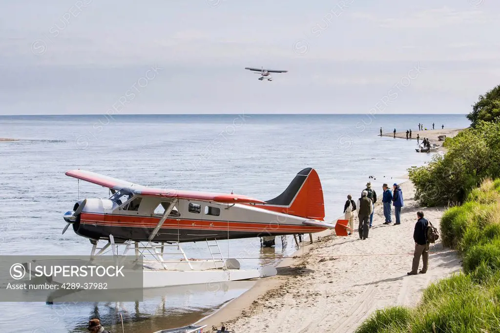 Floatplane Tied To The Sandy Shore Picking Up And Dropping Off Guests With Fishermen And Another Plane In Flight Complete The Scene Of Sport Fishing L...