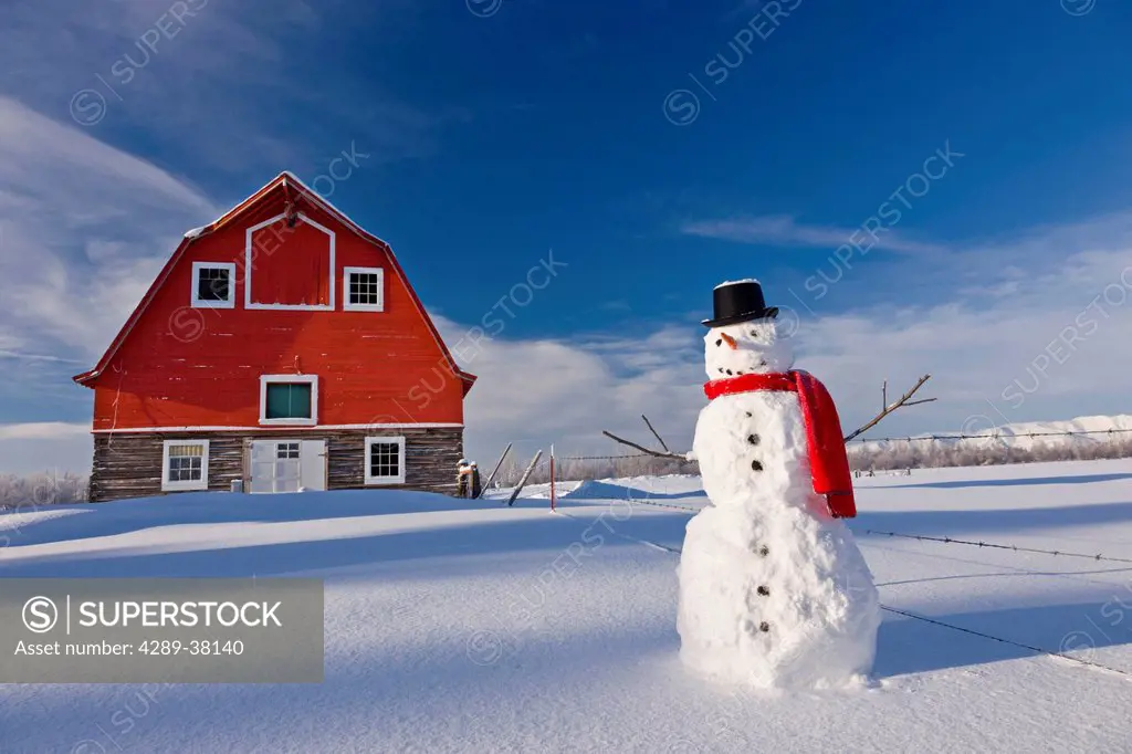 Snowman with red scarf and top hat standing in front of a vintage red barn in winter;Palmer alaska usa