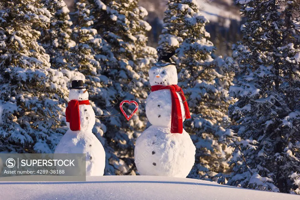 Snowman giving a red heart to a snowwoman and wearing red scarves and black top hats and standing in front of a snowcovered spruce forest;Anchorage al...
