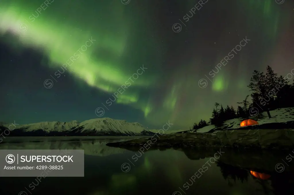 View of the Aurora Borealis (Northern Lights) dancing above the Chugach Mountains with a backpacking tent reflecting in the waters of Turnagain Arm, K...
