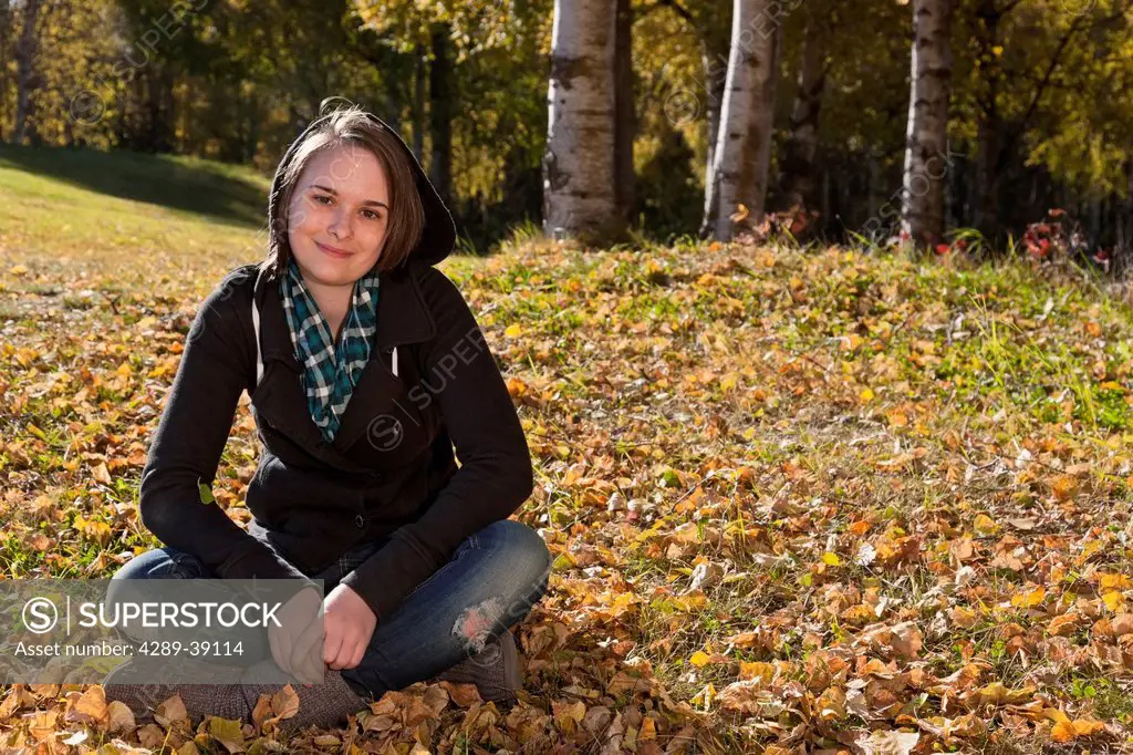 Young woman sitting in the grass with fallen leaves around her, Russian Jack Springs Park, Fall, Anchorage, Southcentral Alaska, USA.
