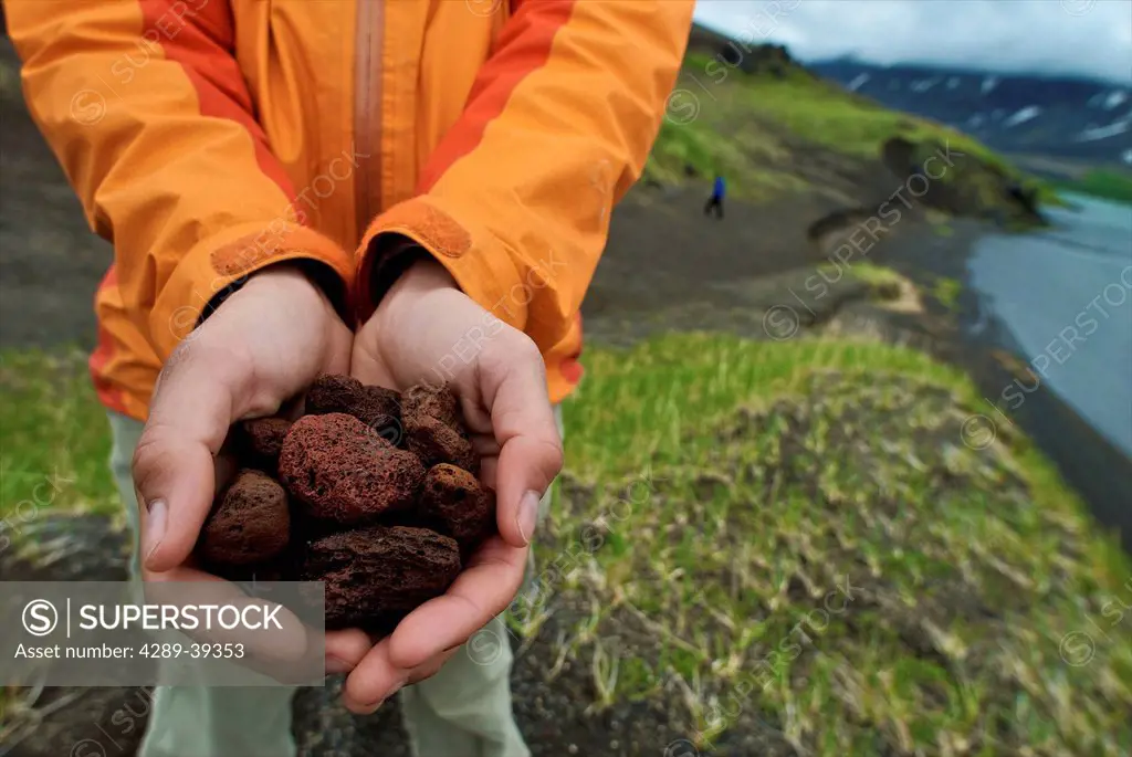 Person Holds Up A Handful Of Lava Rock For Display With Surprise Lake In The Background At Aniakchak National Monument And Preserve In Southwest Alask...