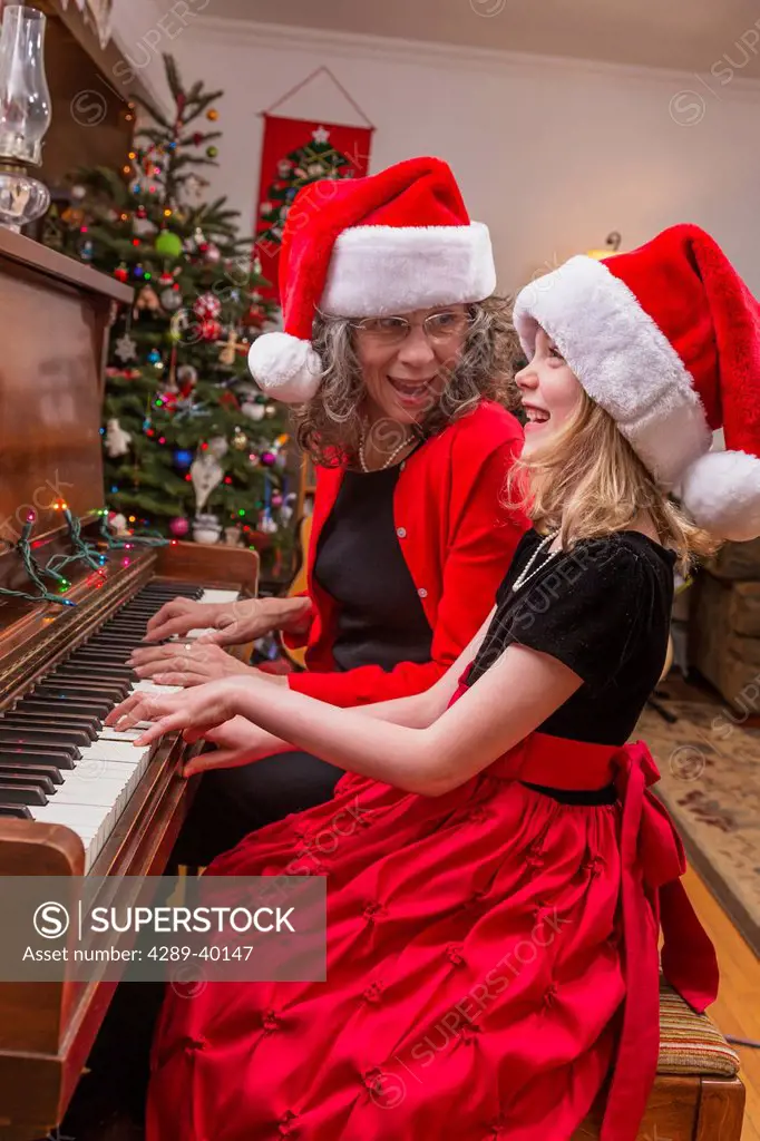 A six year old girl and an older woman both wearing Santa hats playing the piano together, formal dress, house decorated for Christmas.