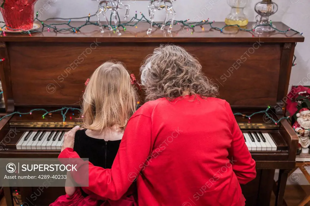A six year old girl and an older woman playing the piano together in formal dress with house decorated for Christmas.