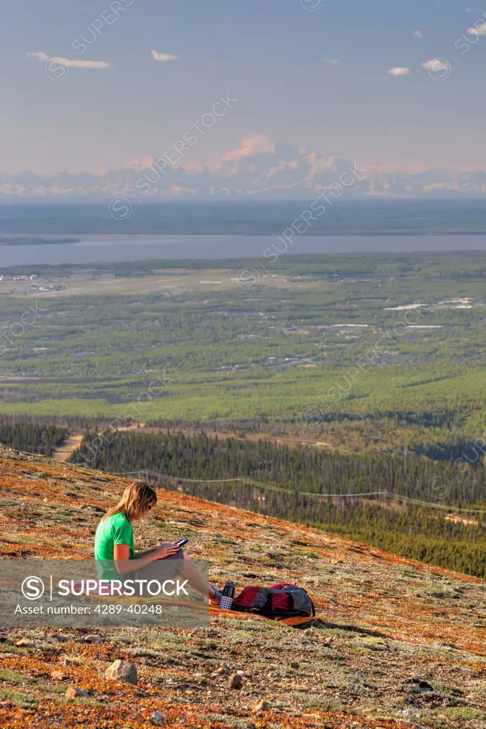 Woman looking at a tablet computer on Blueberry Hill with Denali (Mt. McKinley) in the distance, Chugach State Park, Anchorage, Southcentral Alaska, S...