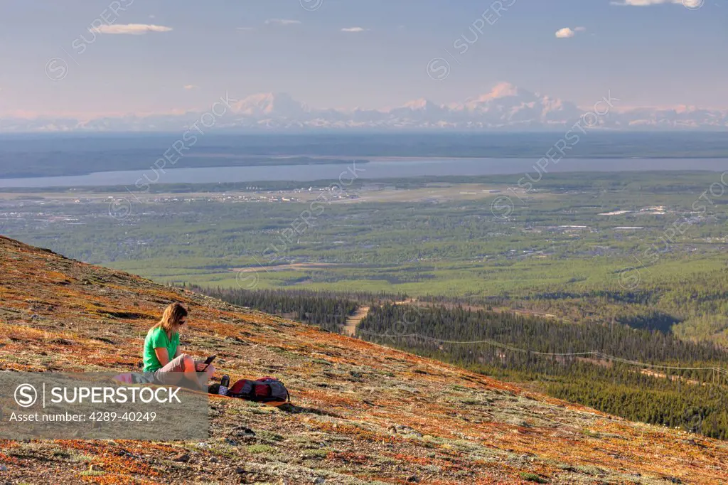Woman looking at a tablet computer on Blueberry Hill with Denali (Mt. McKinley) in the distance, Chugach State Park, Anchorage, Southcentral Alaska, S...