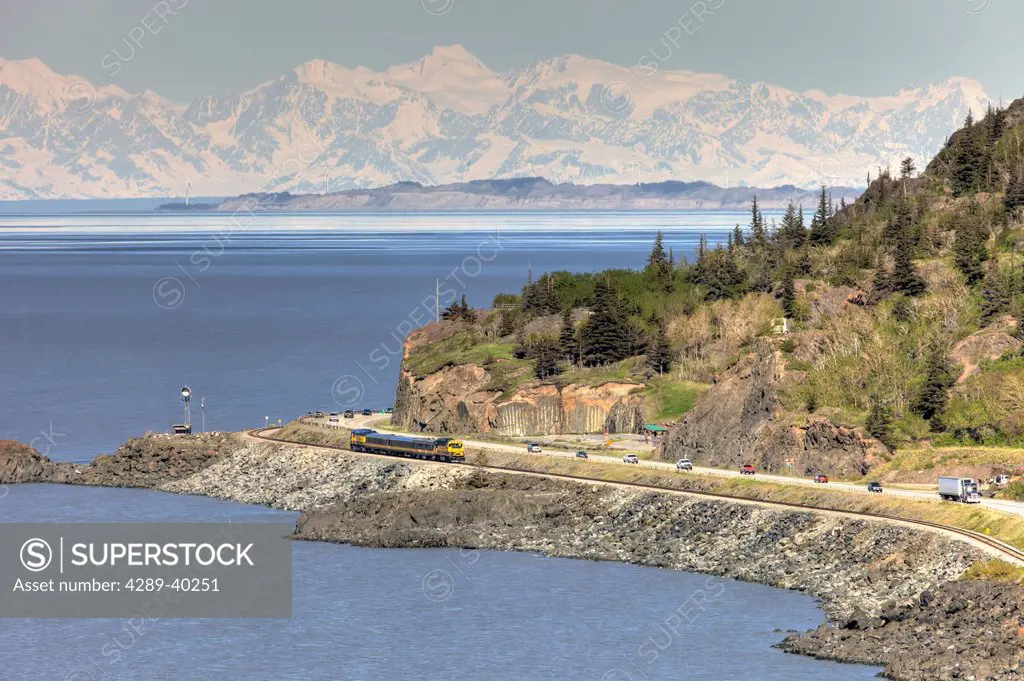 An Alaska Railroad passenger train travels along Turnagain Arm passing traffic on the Seward Highway south of Anchorage with the Alaska Range in the b...