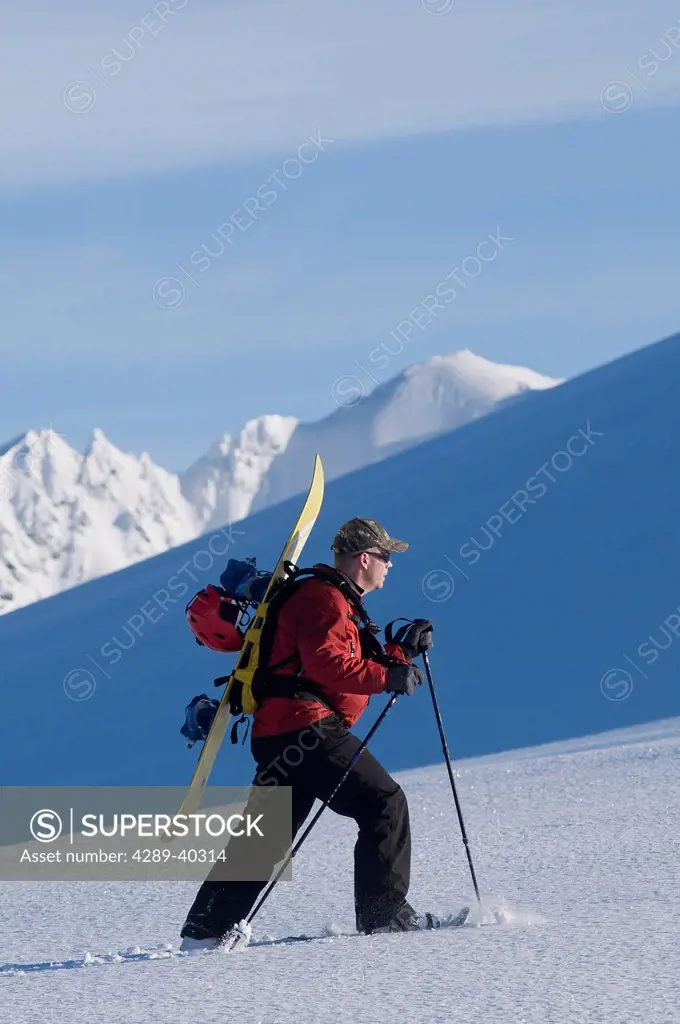Backcountry Snowboarder Hiking Across A Snow Field On The Eagle Glacier With A Snowboard And Helmet Strapped To His Pack, Chugach Mountains, Southcent...