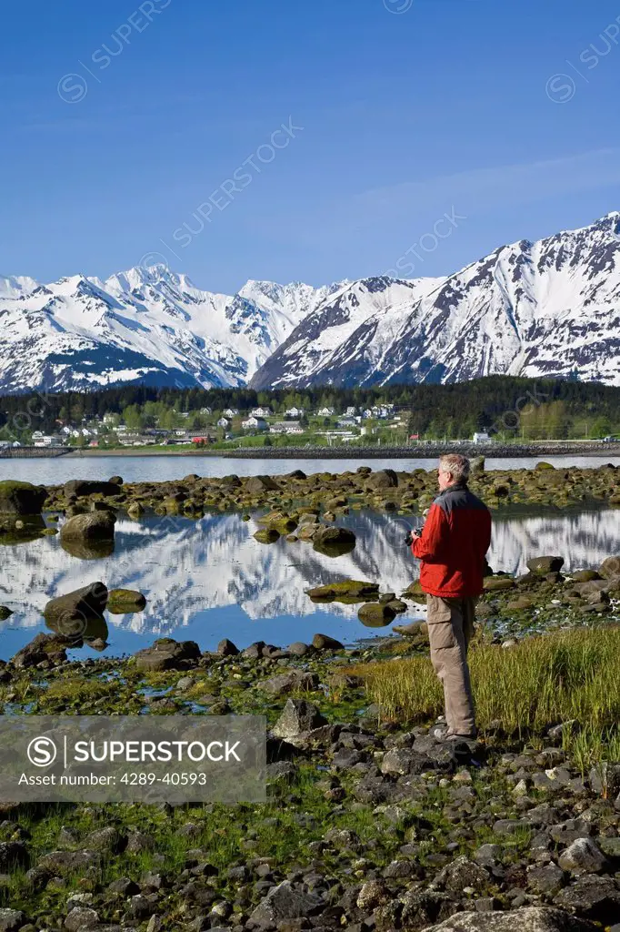 Man Standing Near Shoreline Viewing The Community Of Haines In The Distant. Summer In Southeast Alaska.